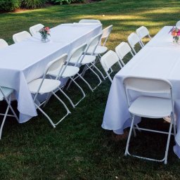 Fanfara Events - Dwa białe stoły nakryte białymi obrusami i otoczone białymi krzesłami na trawniku, na stołach wazony z kwiatami, przygotowane na imprezę plenerową.
