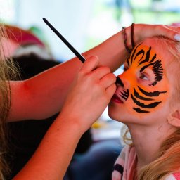 Fanfara Events - Animator maluje twarz dziewczynki w tygrysa cienkim pędzlem, widoczny fragment pomarańczowych i czarnych pasków oraz fragment różowej bluzki dziewczynki.