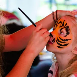 Animator maluje twarz dziewczynki w tygrysa cienkim pędzlem, widoczny fragment pomarańczowych i czarnych pasków oraz fragment różowej bluzki dziewczynki.