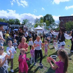 Fanfara Events - Animator w stroju pirata puszcza duże bańki mydlane na trawie, wokół zgromadzone dzieci obserwują i próbują je dotknąć, w tle widoczny tłum ludzi i budynek.