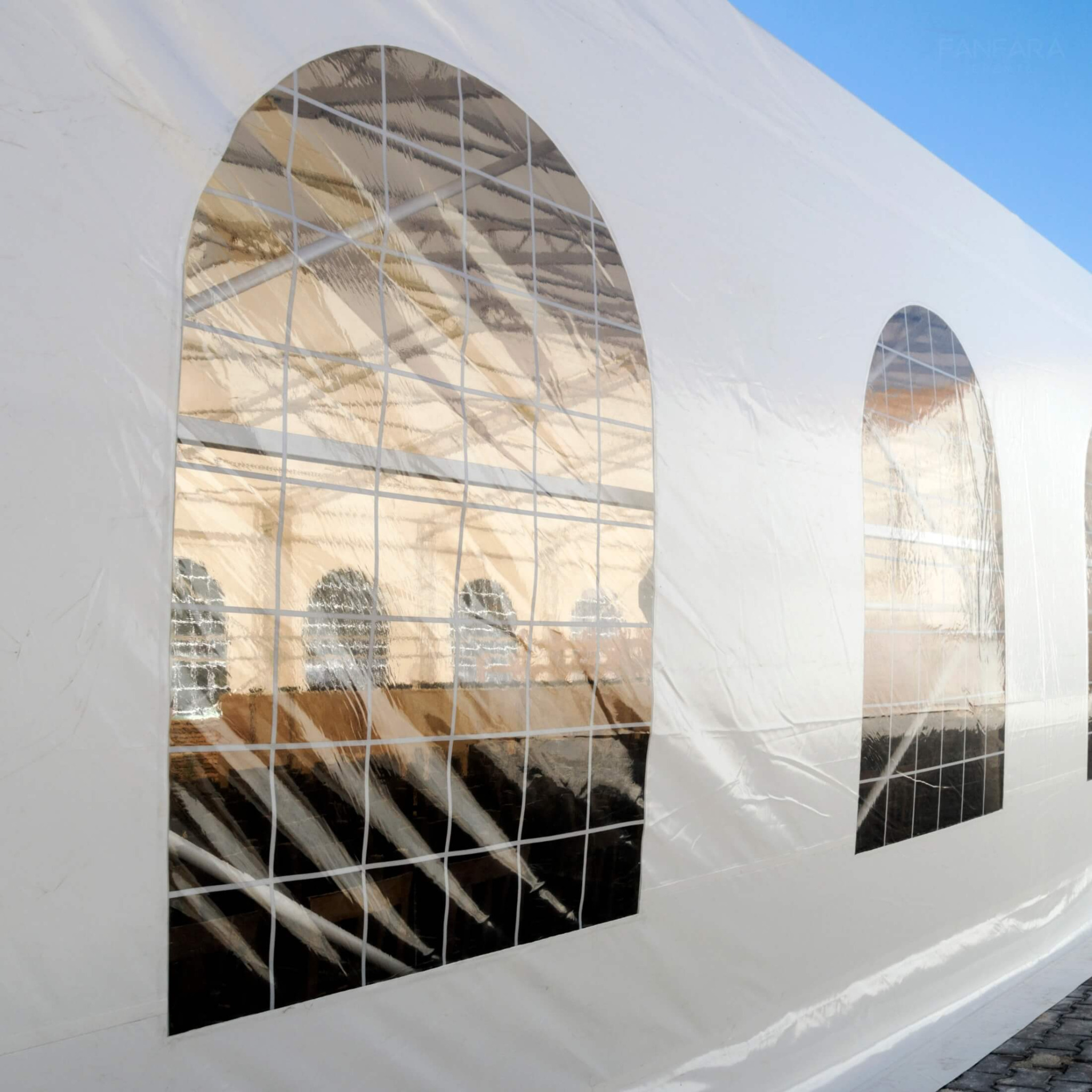 Biały namiot eventowy z przezroczystymi, łukowatymi oknami, przez które widać konstrukcję wewnątrz, na tle błękitnego nieba.