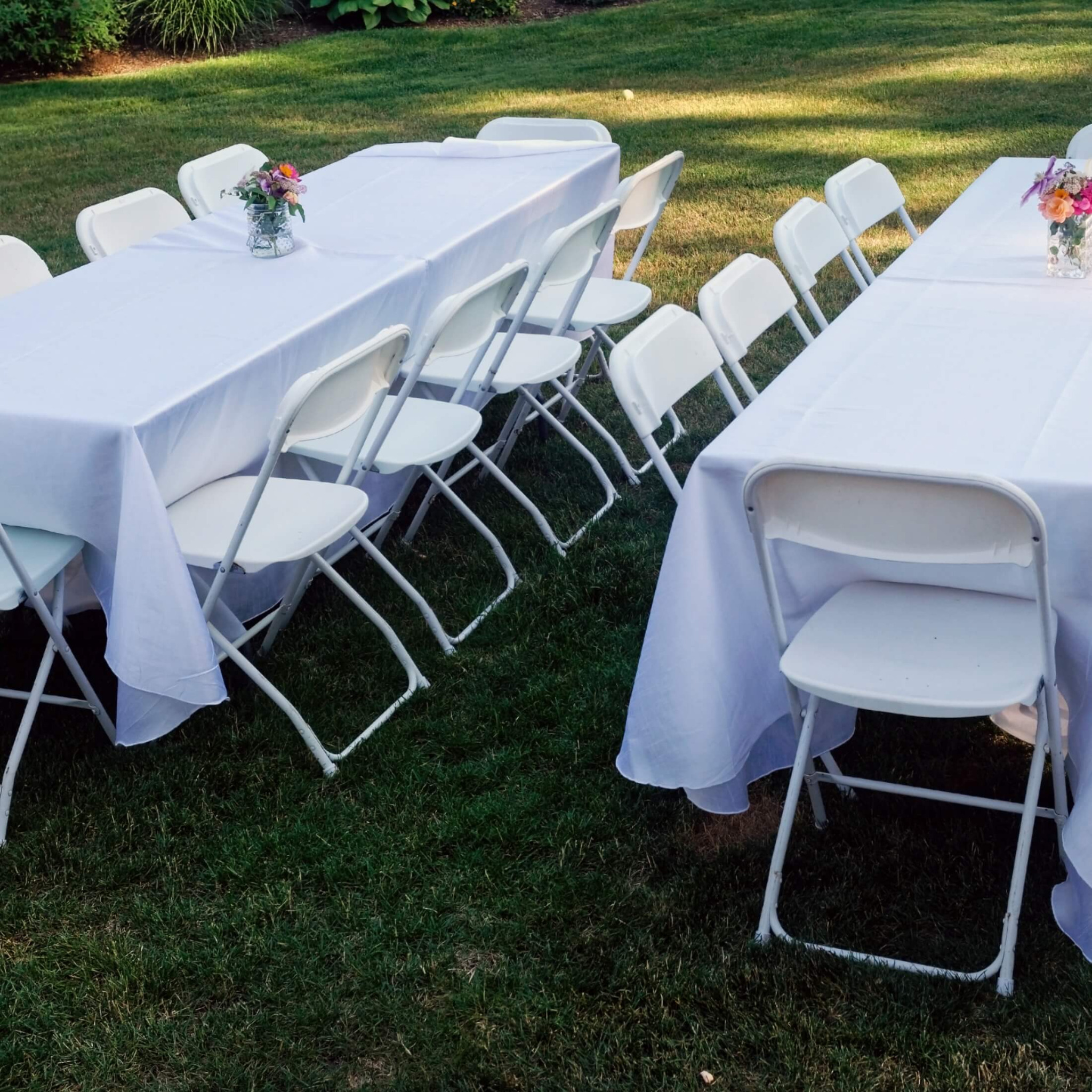 Dwa białe stoły nakryte białymi obrusami i otoczone białymi krzesłami na trawniku, na stołach wazony z kwiatami, przygotowane na imprezę plenerową.