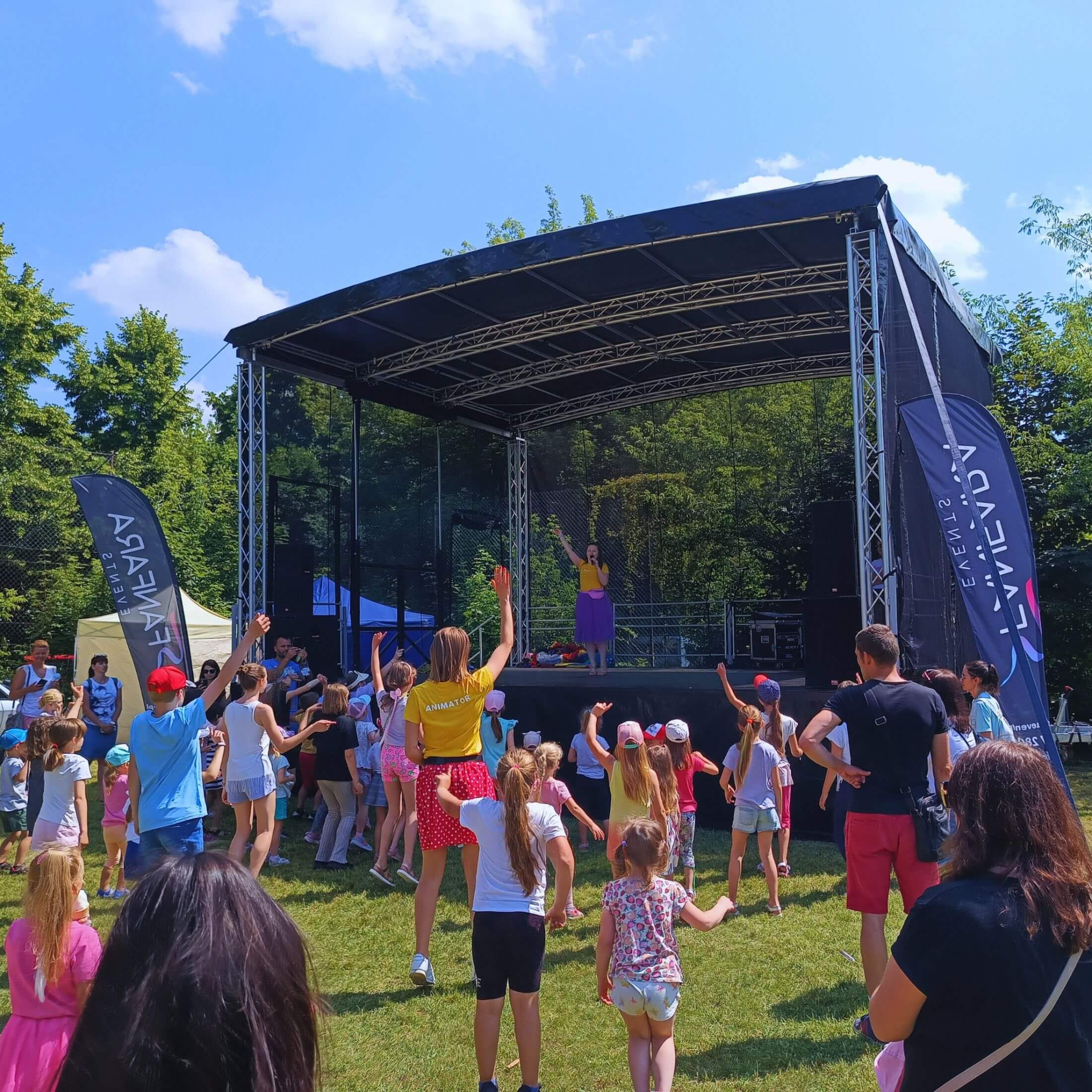 Grupa dzieci tańczących na trawie przed sceną plenerową, na której występuje kobieta w fioletowej spódnicy i żółtej bluzce, z boku widoczny baner reklamowy.