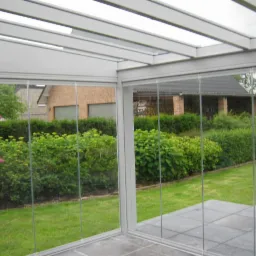 Szklana zabudowa tarasu z widokiem na zadbany ogród i ceglany budynek w tle, szara konstrukcja ramy i zadaszenia, płytki tarasowe na podłodze.