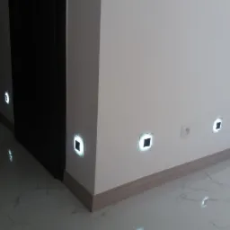 Świecące kwadratowe lampki podłogowe wpuszczone w ścianę, oświetlające marmurową posadzkę wzdłuż ściany i drzwi.