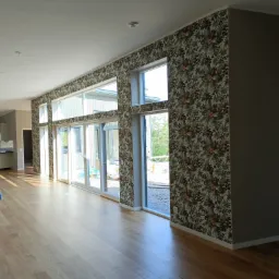 Jasne wnętrze domu z drewnianą podłogą i ścianą w tropikalne wzory, duże okna przesuwne wpuszczają dużo światła.