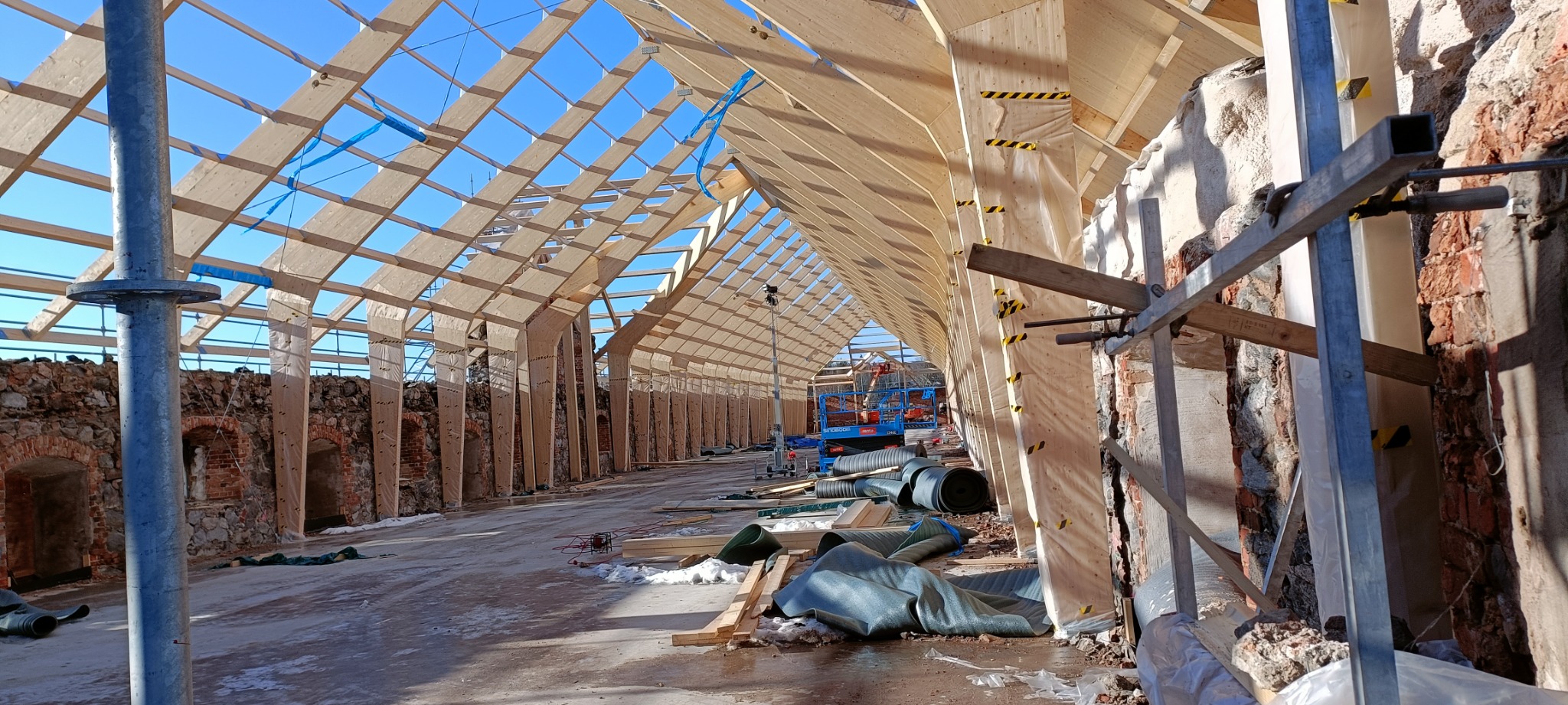 Wnętrze w trakcie budowy domu szkieletowego z widoczną konstrukcją drewnianą dachu, zabezpieczonymi ścianami z cegły i podnośnikiem hydraulicznym w tle.