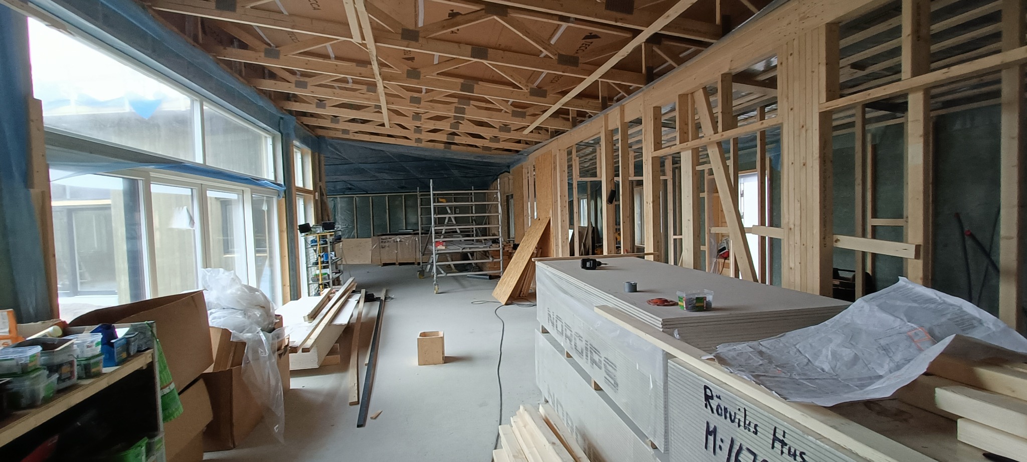 Wnętrze w trakcie budowy: widoczne drewniane elementy konstrukcyjne ścian i stropu, duże okna z niebieską folią ochronną, rusztowanie oraz stos płyt gipsowo-kartonowych z planem budowy na wierzchu.