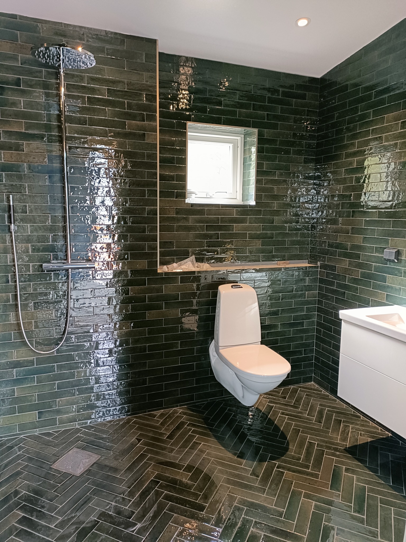 Nowoczesna łazienka z ciemnozielonymi, błyszczącymi płytkami ułożonymi w cegiełkę na ścianach i jodełkę na podłodze, biała wisząca toaleta i umywalka, chromowana deszczownica.