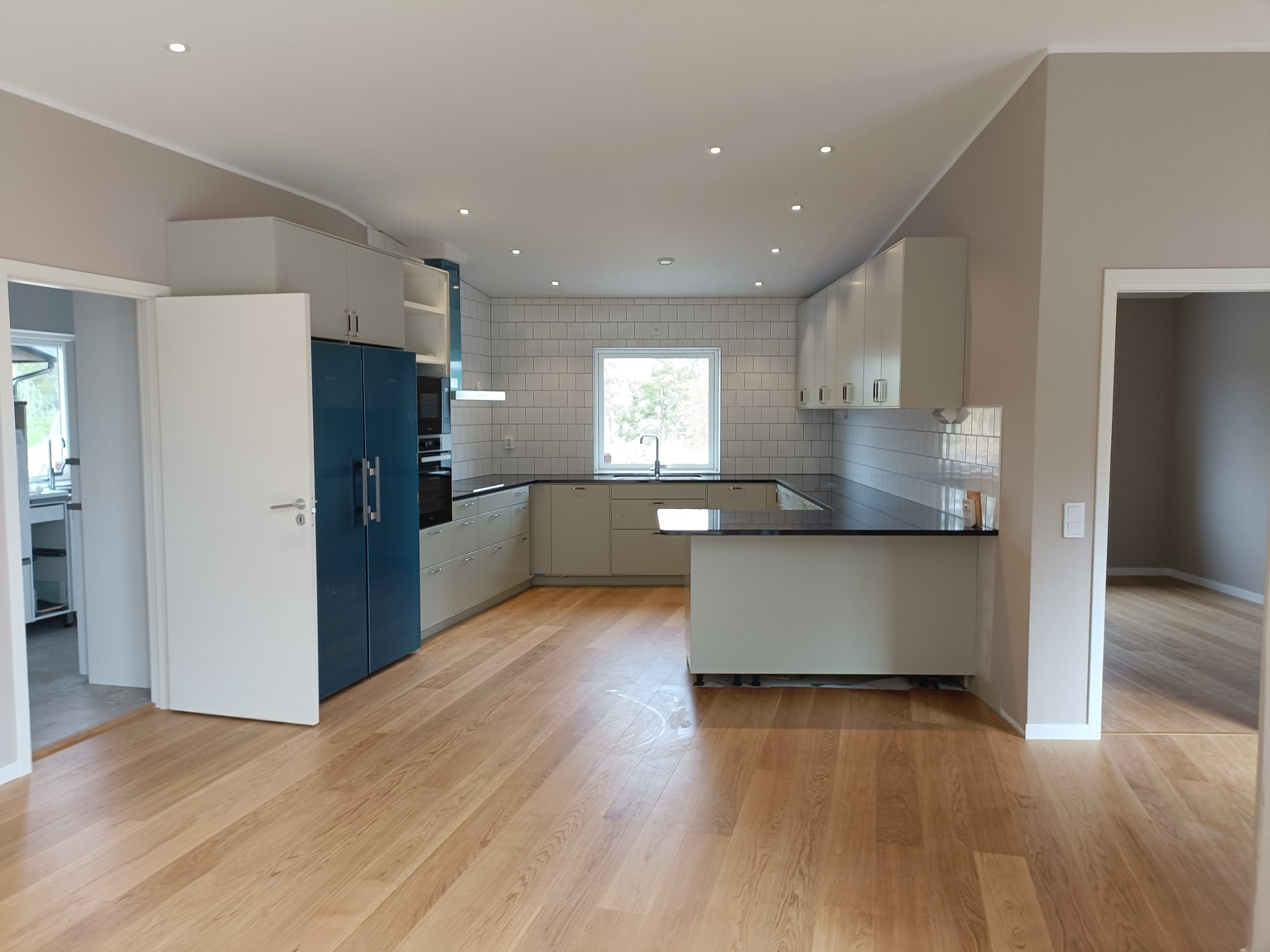 Jasna kuchnia z drewnianą podłogą, białą glazurą na ścianie i niebieską lodówką. Widok na okno z widokiem na drzewa.