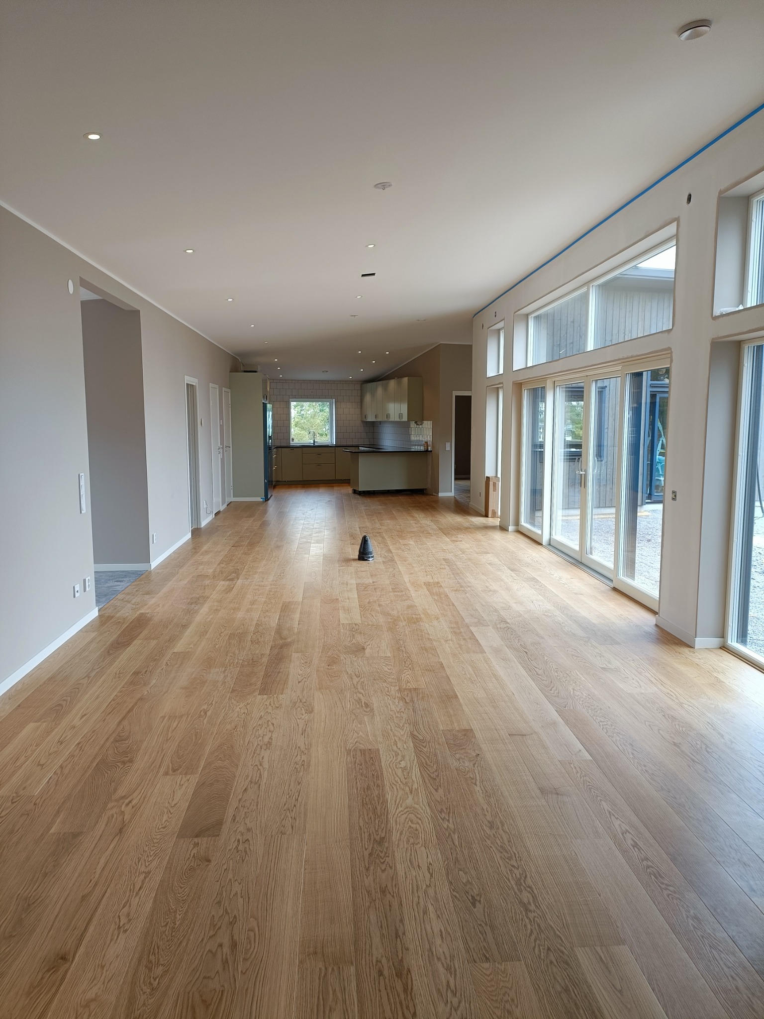 Jasne, przestronne wnętrze domu z drewnianą podłogą, dużymi oknami i widokiem na kuchnię w tle. Niebieska taśma malarska wzdłuż sufitu nad oknami.