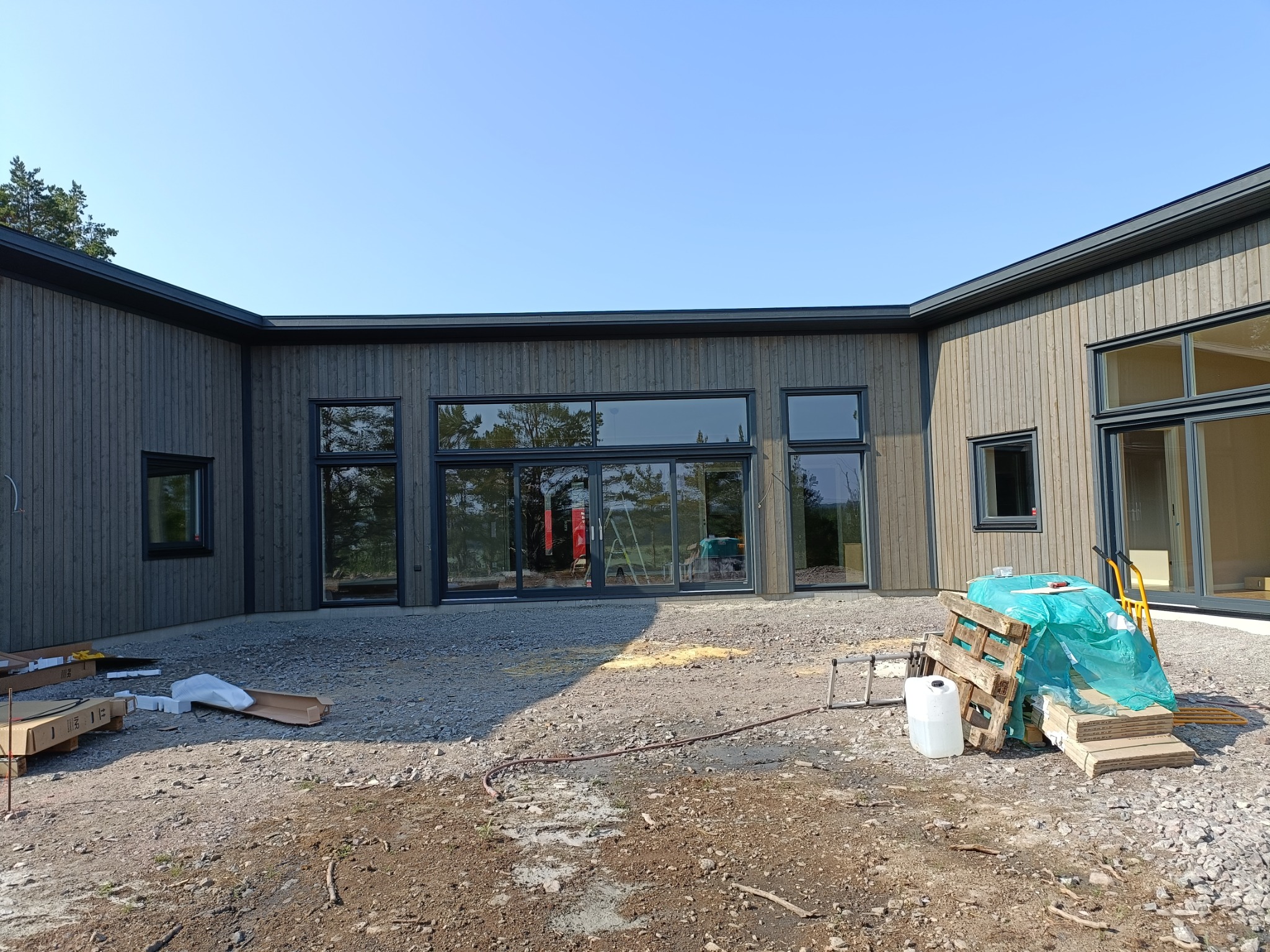 Surowy stan budowy: Zewnętrzny widok na dom z szarej deski z dużymi oknami, widoczne materiały budowlane, palety i narzędzia na żwirowym podłożu przed budynkiem, błękitne niebo w tle.