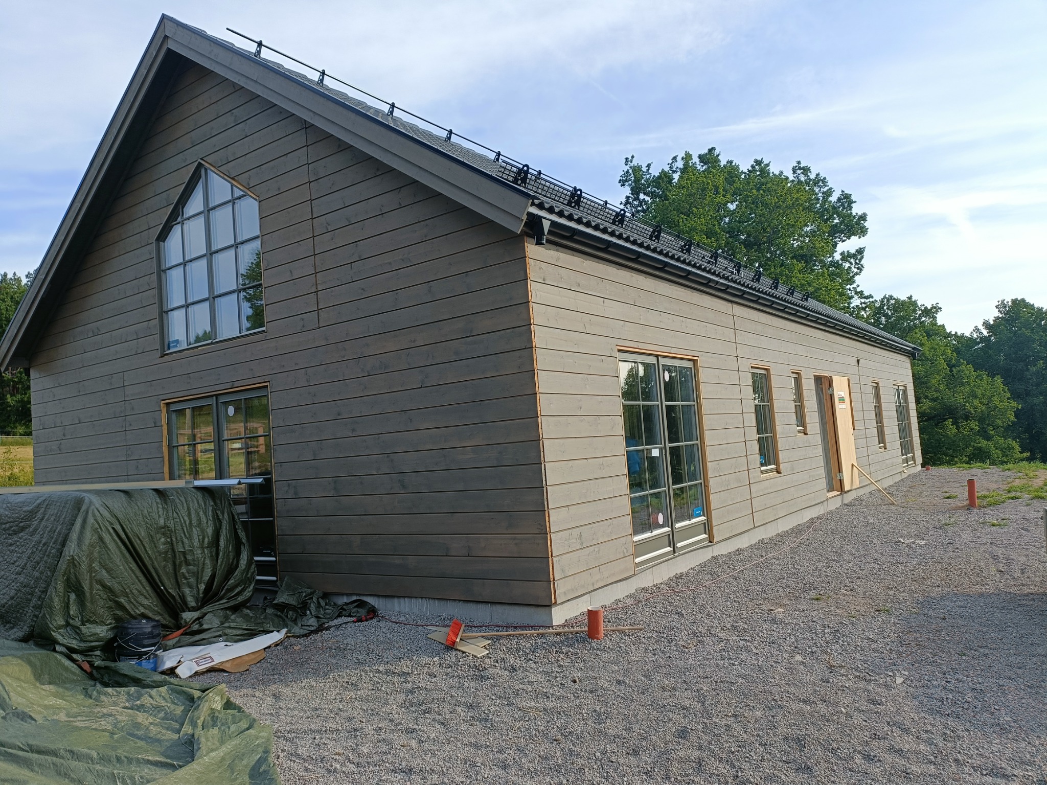 Nowoczesny dom o prostej bryle z drewnianą elewacją w odcieniach szarości, z charakterystycznym trójkątnym oknem na szczycie, częściowo zakryty plandeką i materiałami budowlanymi na żwirowym podłożu.