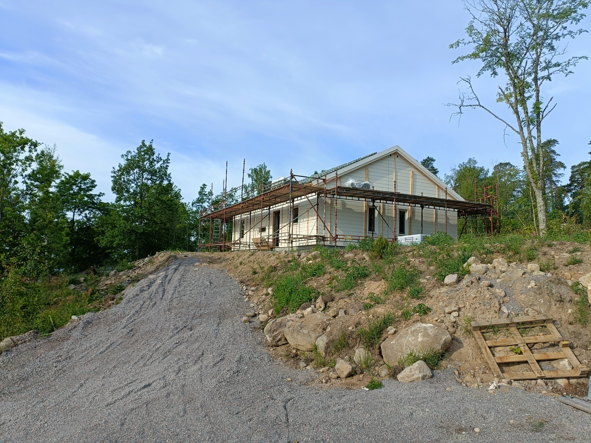 Niedokończony dom szkieletowy z jasną elewacją, otoczony rusztowaniem, widoczny podjazd z szutru i drewniana paleta na zboczu.