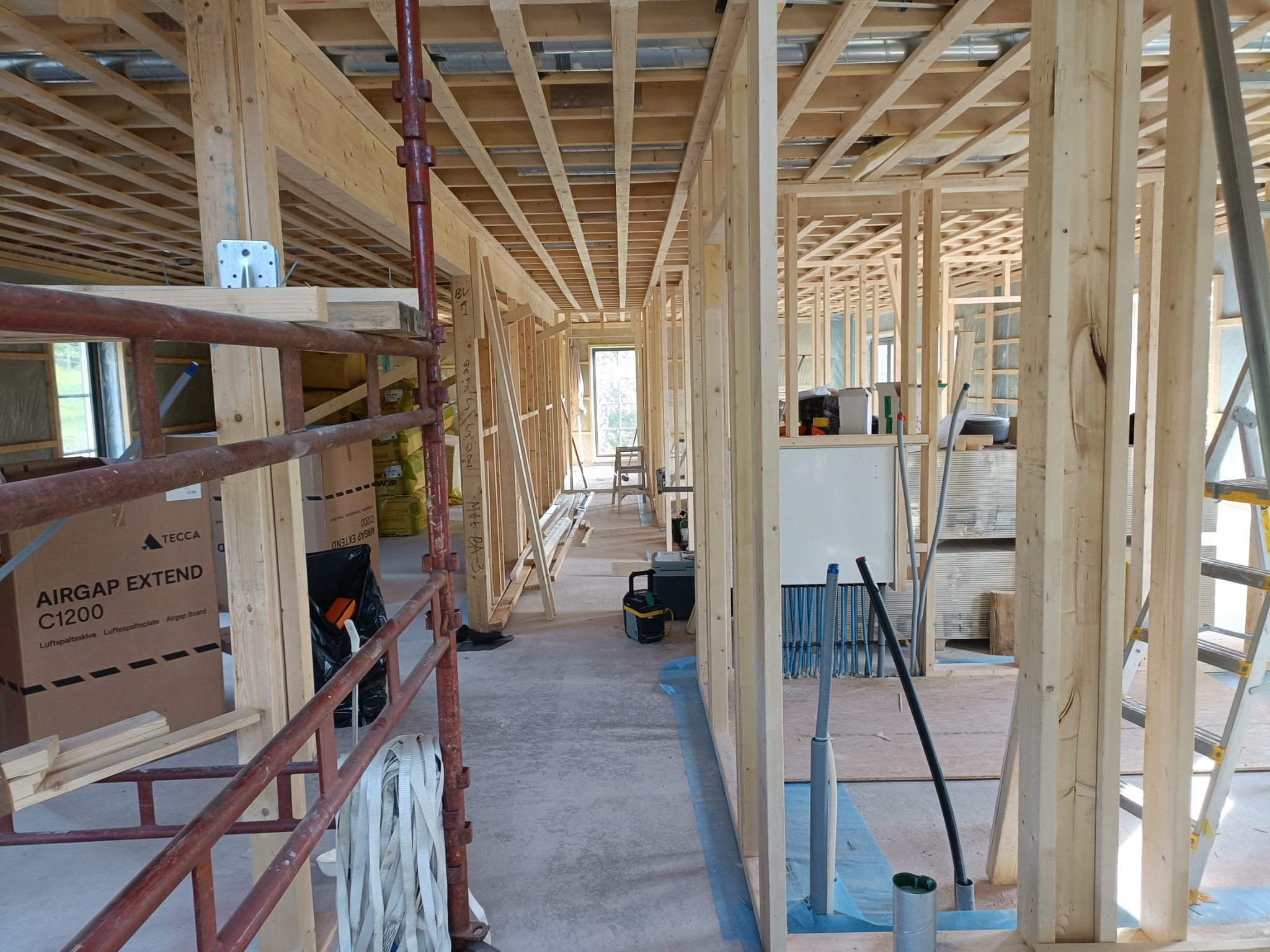 Wnętrze domu w trakcie budowy, widoczna drewniana konstrukcja szkieletowa, rusztowania, materiały budowlane i narzędzia. Przez otwór w ścianie widać dalszą część budynku i okno.