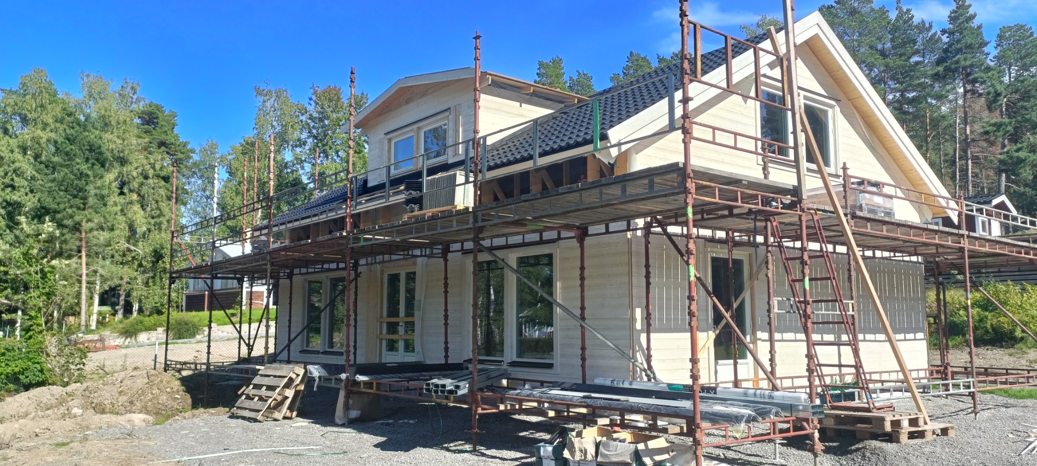 Wznoszony dwukondygnacyjny dom z jasnego drewna, otoczony rusztowaniami i zielenią, z widocznym fragmentem dachu pokrytego ciemną dachówką. Na pierwszym planie widoczne materiały budowlane i palety.