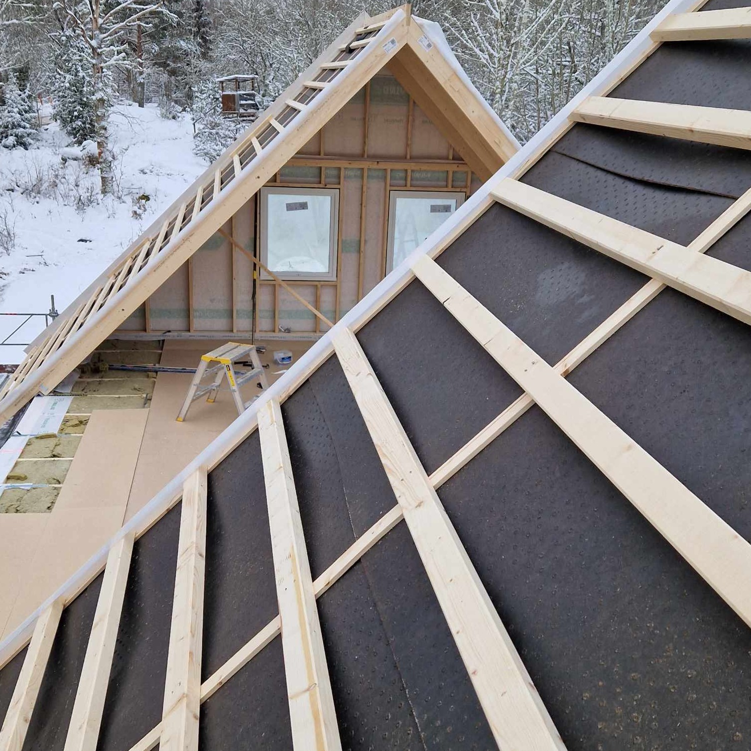 Wznoszenie konstrukcji domu w zimowym krajobrazie: widok na szkielet dachu z czarną membraną i fragment ściany z oknami, drabina w tle.