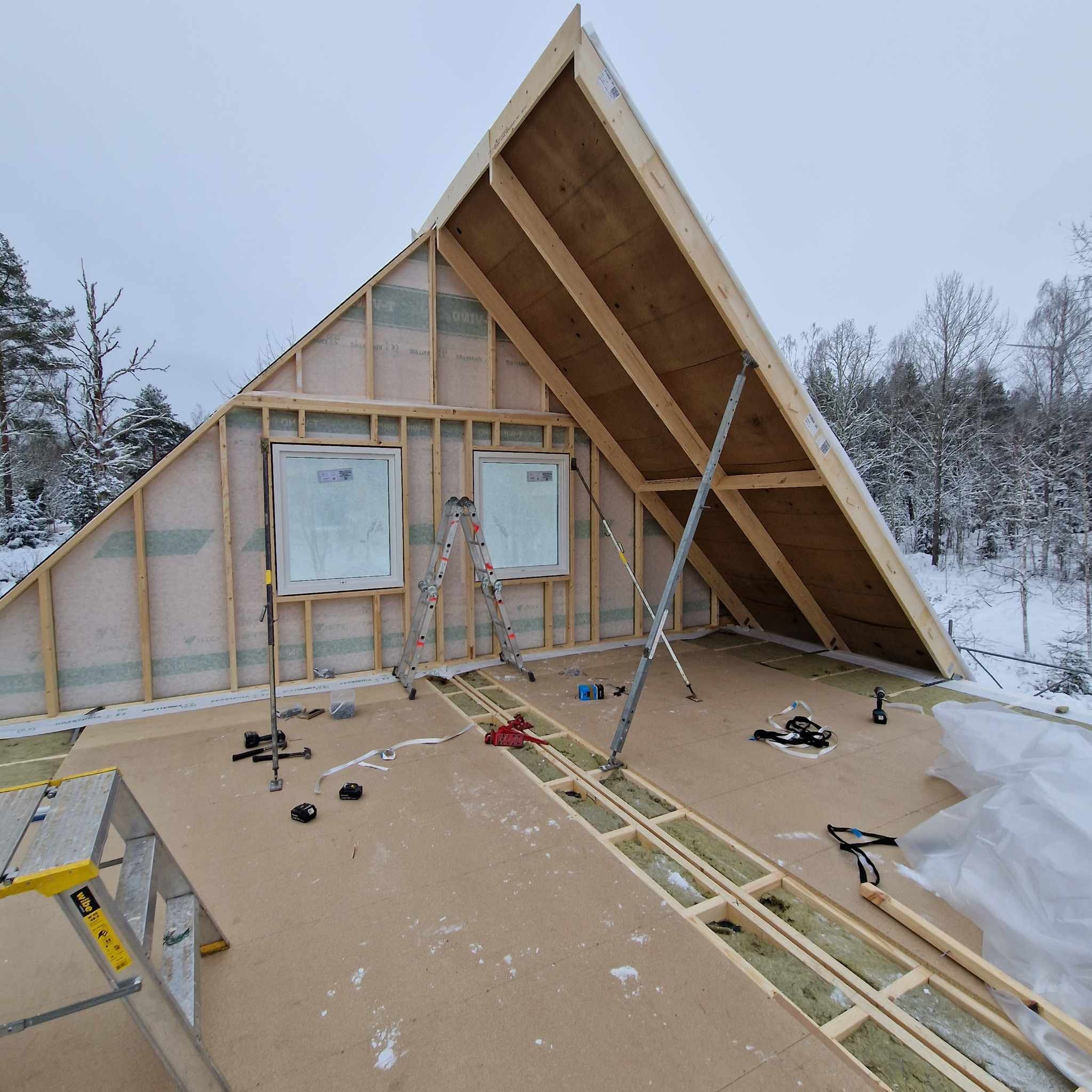 Wnętrze w trakcie budowy z widoczną drewnianą konstrukcją dachu, ścian z oknami oraz izolacją w podłodze, na tle zimowego krajobrazu.