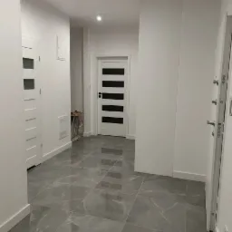 Korytarz wykończony w szaro-białej kolorystyce z marmurową posadzką, widoczne trzy białe drzwi z różnymi wzorami, oświetlenie punktowe sufitowe.