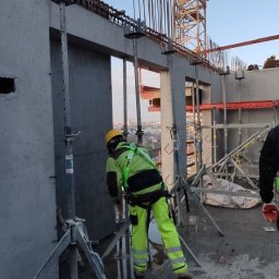 IS BUD® Mykola Mostovyi - Wysoka kondygnacja budynku w trakcie budowy. Widoczne szalunki, stalowe podpory i pracownicy w kaskach analizujący dokumentację na tle panoramy miasta.