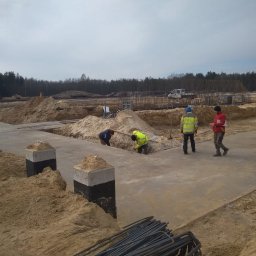 IS BUD® Mykola Mostovyi - Prace zbrojarskie na budowie: robotnicy w odblaskowych ubraniach układają stal zbrojeniową w wykopach pod fundamenty. Widoczne betonowe słupy i przygotowany teren.
