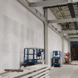 IS BUD® Mykola Mostovyi - Wnętrze hali w trakcie prac wykończeniowych: ściany po gładzi, stalowe konstrukcje pod sufitem, podnośniki koszowe i elementy konstrukcyjne w pakietach.