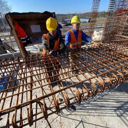 Dwóch pracowników w kaskach i kamizelkach układa zbrojenie na budowie, widok z góry, w tle autobus i drzewa.