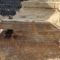 Szeroki kadr z placu budowy: zbrojenie fundamentów z widocznym wykopem, zabezpieczeniem ścian wykopu i sprzętem budowlanym. Widok z góry na siatkę zbrojeniową.