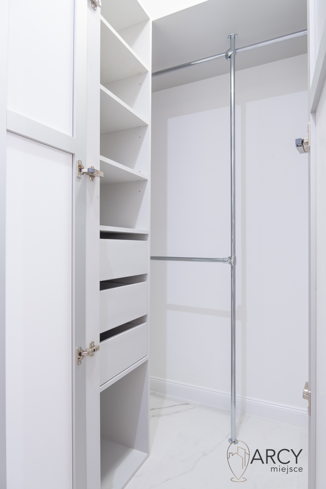 Wnętrze białej, otwartej garderoby z półkami, szufladami i metalowym drążkiem na ubrania, wykończonej marmurową podłogą, widoczny zawias drzwiowy.