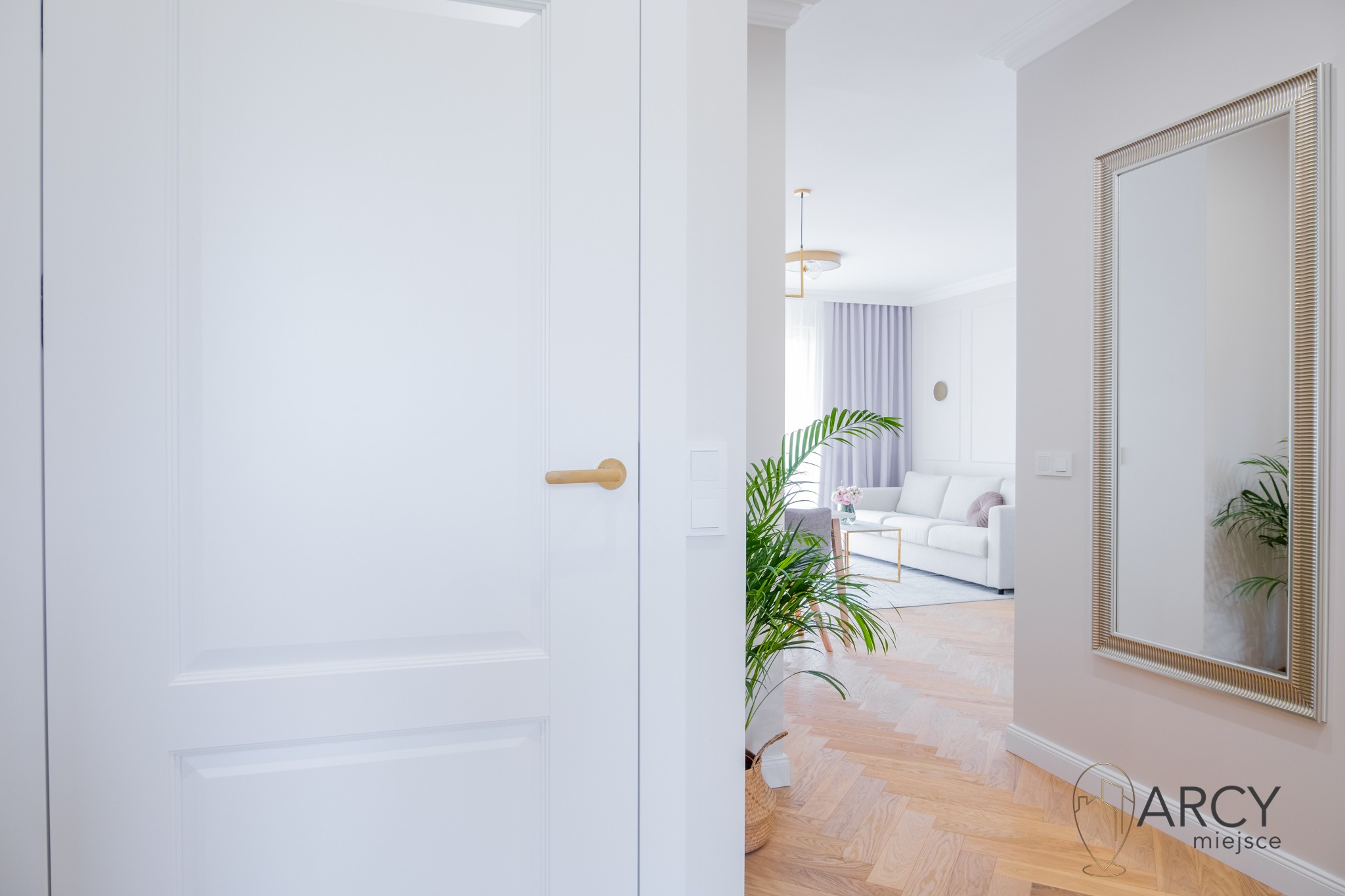 Jasne wnętrze mieszkania z parkietem w jodełkę, białe drzwi z złotą klamką i widok na salon z sofą i lustrem w ozdobnej ramie.