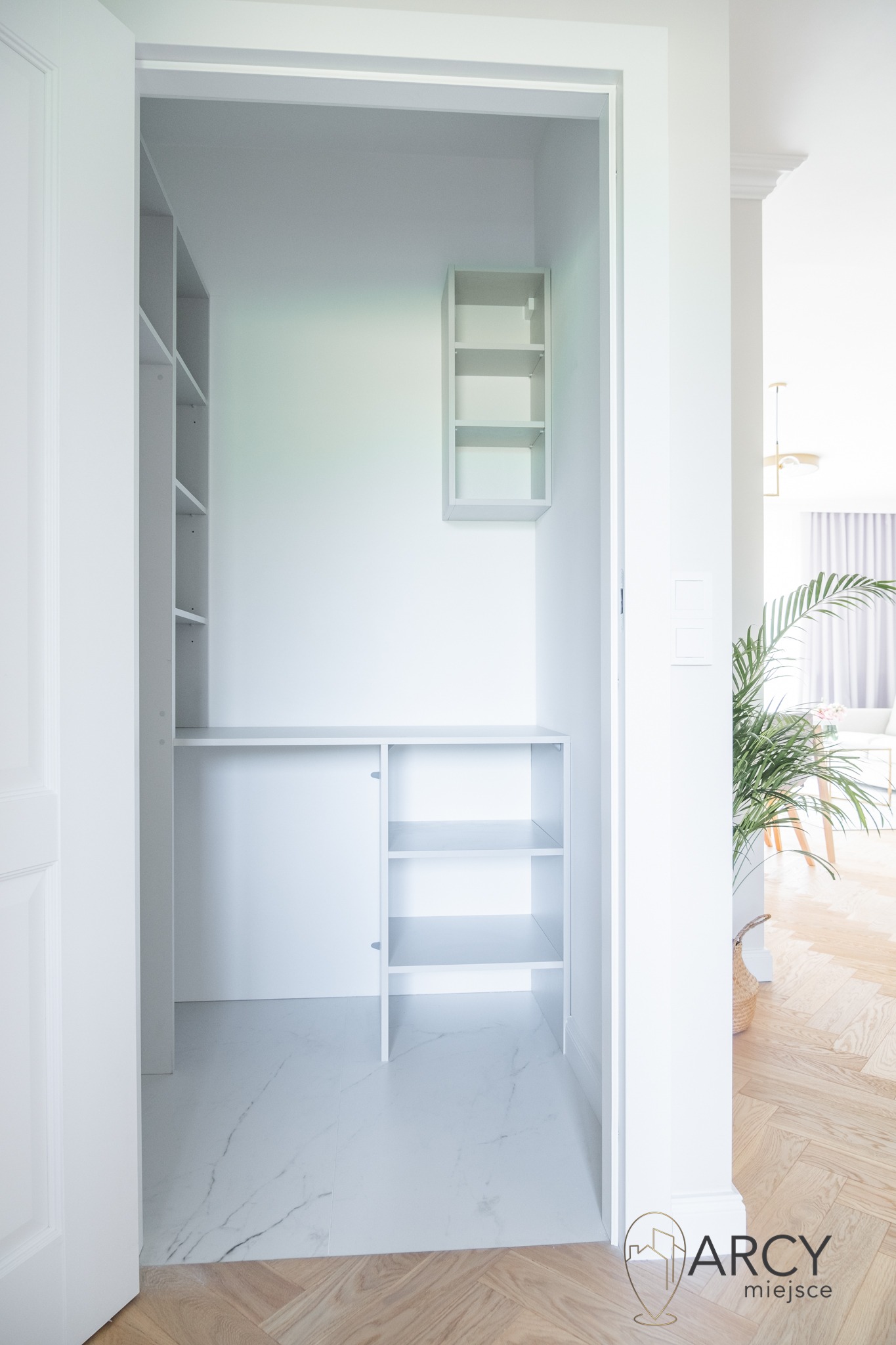 Wnętrze garderoby z białymi półkami i blatem, marmurowe płytki na podłodze, widok z korytarza z parkietem w jodełkę i rośliną doniczkową w tle.