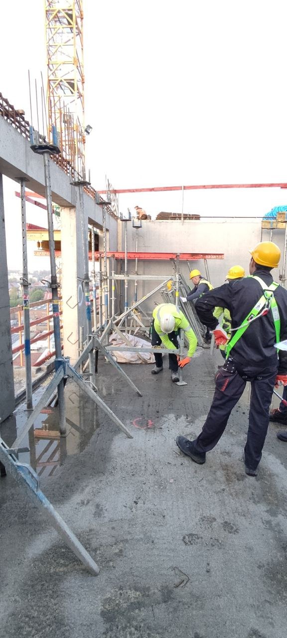 Ekipa budowlana w kaskach i kamizelkach pracuje na budowie, montując szalunki na betonowym stropie. W tle widoczny żuraw budowlany i elementy konstrukcji.