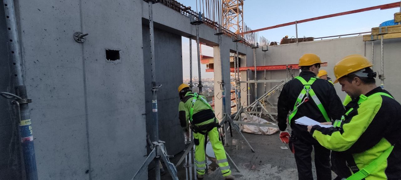 Wysoka kondygnacja budynku w trakcie budowy. Widoczne szalunki, stalowe podpory i pracownicy w kaskach analizujący dokumentację na tle panoramy miasta.