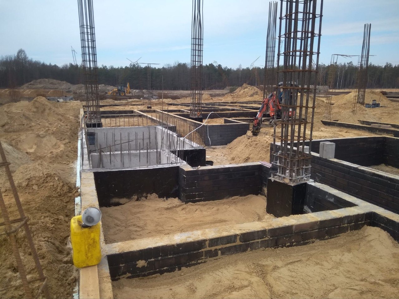 Fundamenty domu w trakcie budowy: zbrojenie, bloczki fundamentowe, izolacja. W tle koparka i hałdy piachu.