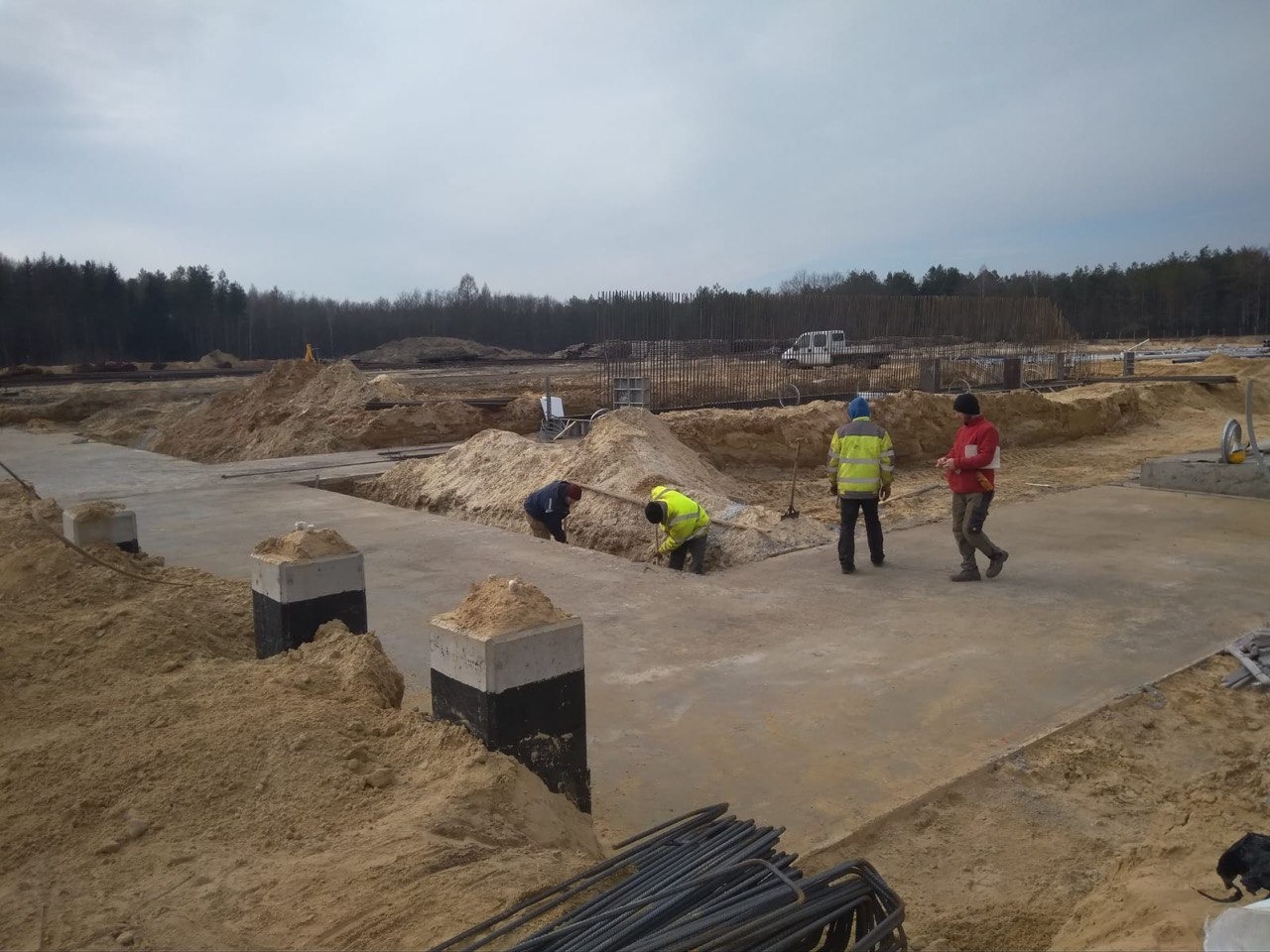 Prace zbrojarskie na budowie: robotnicy w odblaskowych ubraniach układają stal zbrojeniową w wykopach pod fundamenty. Widoczne betonowe słupy i przygotowany teren.