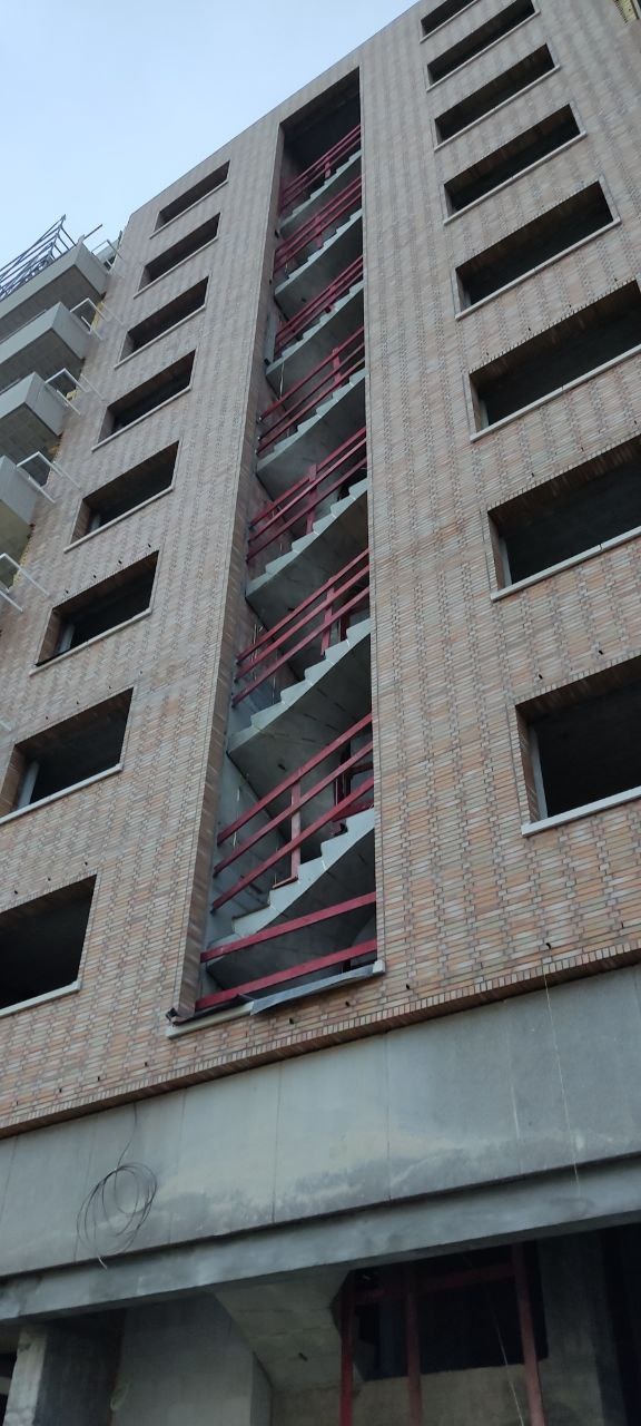 Wysoki budynek w trakcie budowy, widoczne schody z czerwonymi barierkami, ceglana elewacja i niezamontowane okna. Ujęcie z dołu, pochmurne niebo.