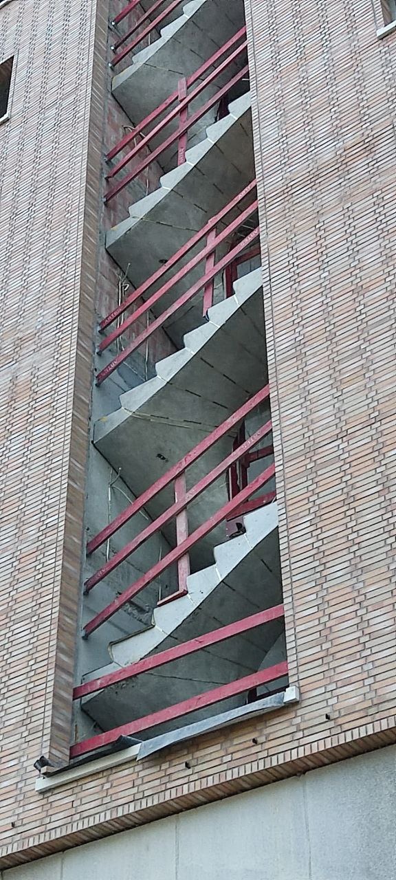 Surowe, betonowe schody w trakcie budowy, z czerwonymi barierkami ochronnymi, widoczne z zewnątrz budynku z ceglaną elewacją. Ujęcie z dołu, podkreślające pionowość konstrukcji.