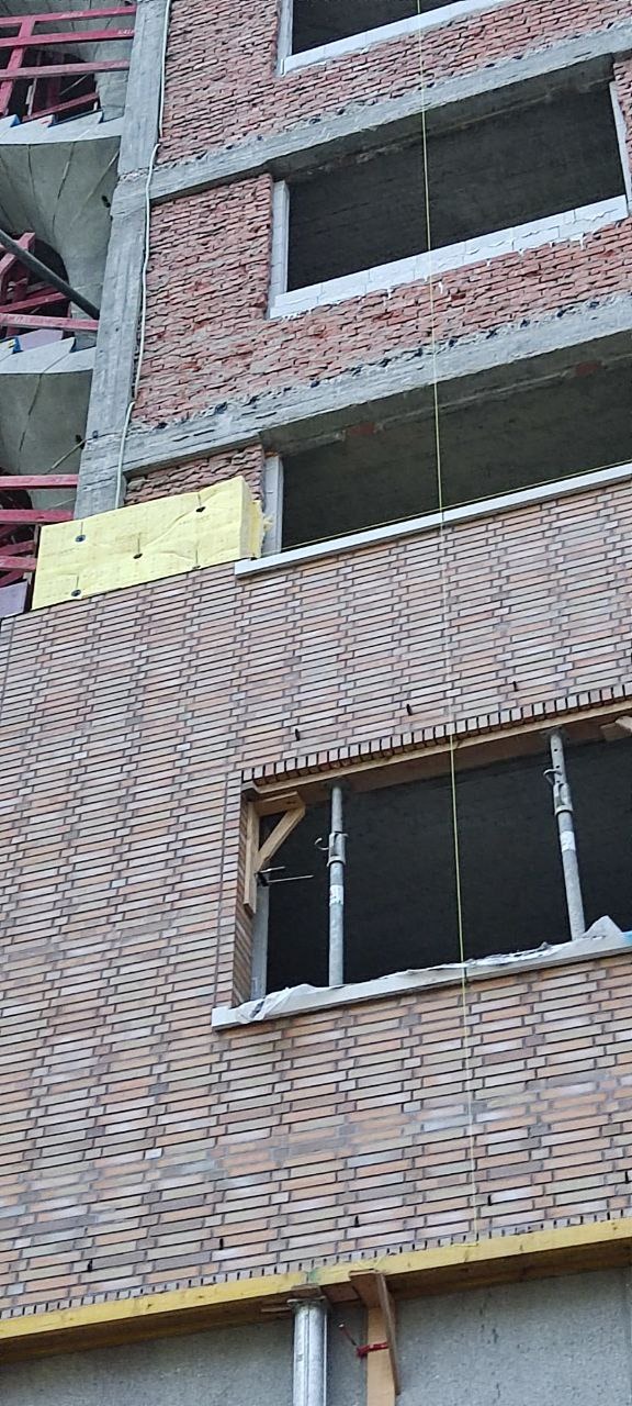 Wznoszenie muru z cegły klinkierowej na budynku w trakcie budowy. Widoczne okna bez ram, podpory i izolacja termiczna. Detal elewacji z cegły.