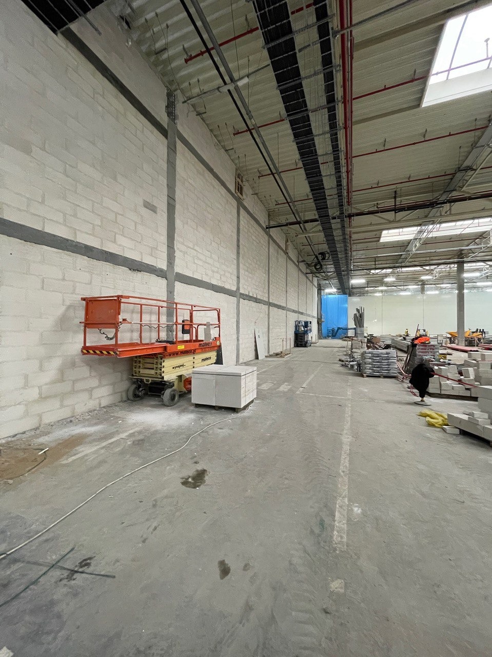 Wnętrze hali w trakcie prac wykończeniowych: ściany z bloków, podnośnik koszowy, materiały budowlane. Widoczne instalacje pod sufitem i oświetlenie.