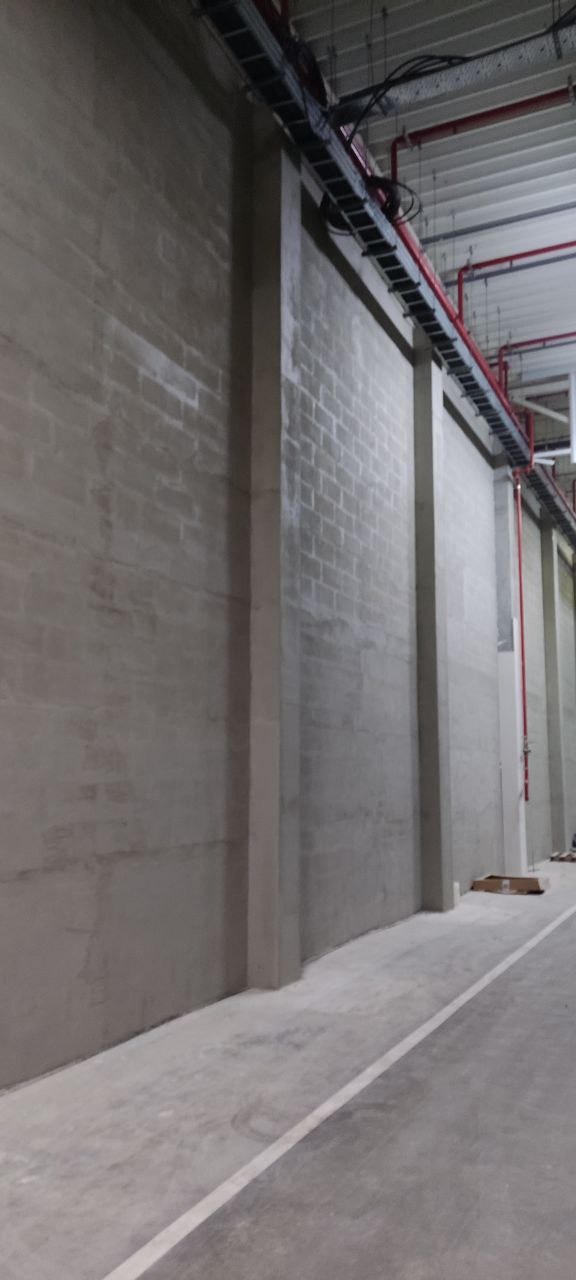 Surowa ściana z betonu komórkowego z pionowymi wzmocnieniami, widoczne instalacje pod sufitem. Minimalistyczne wykończenie hali przemysłowej w szarościach.