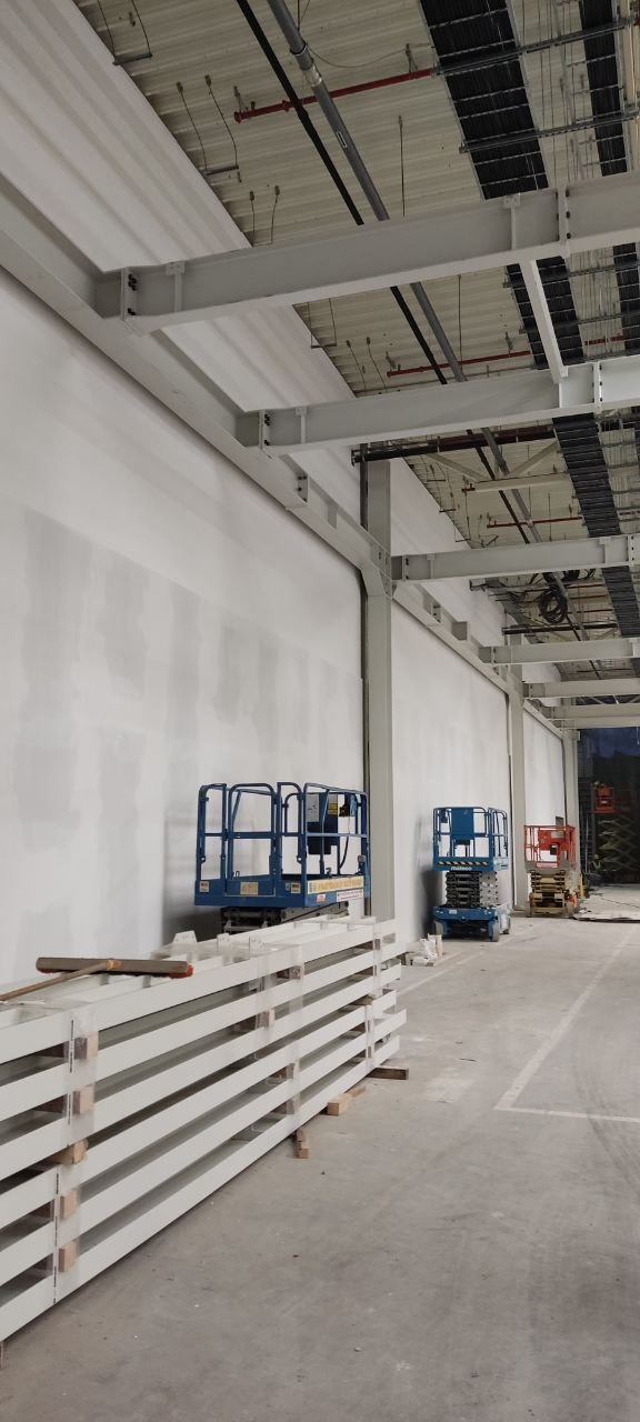 Wnętrze hali w trakcie prac wykończeniowych: ściany po gładzi, stalowe konstrukcje pod sufitem, podnośniki koszowe i elementy konstrukcyjne w pakietach.