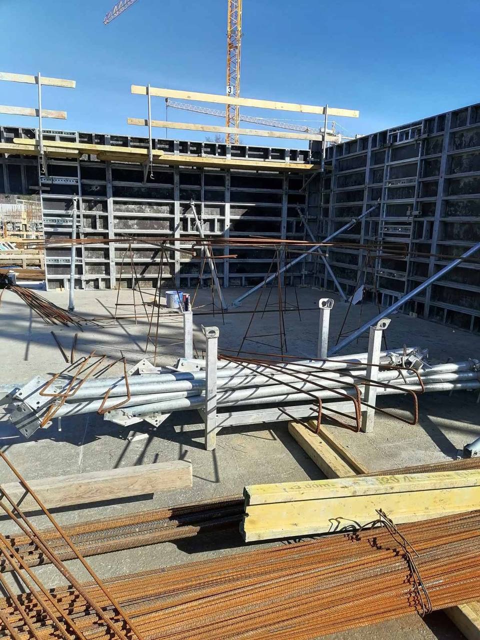 Szare szalunki zbrojeniowe w wykopie, przygotowane do zalania betonem. Na pierwszym planie wiązki prętów zbrojeniowych i podpory. W tle żuraw budowlany.