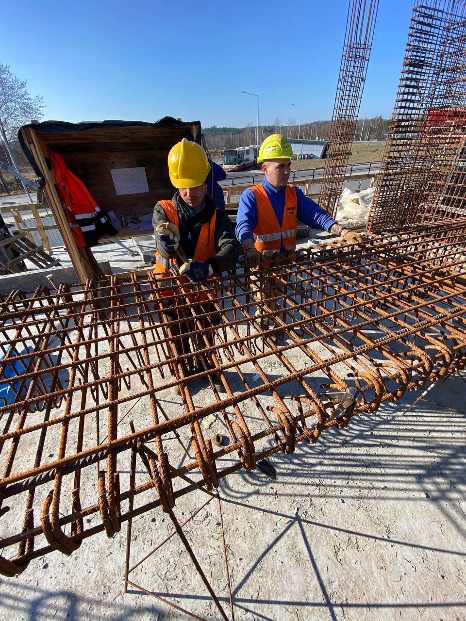 Dwóch pracowników w kaskach i kamizelkach układa zbrojenie na budowie, widok z góry, w tle autobus i drzewa.