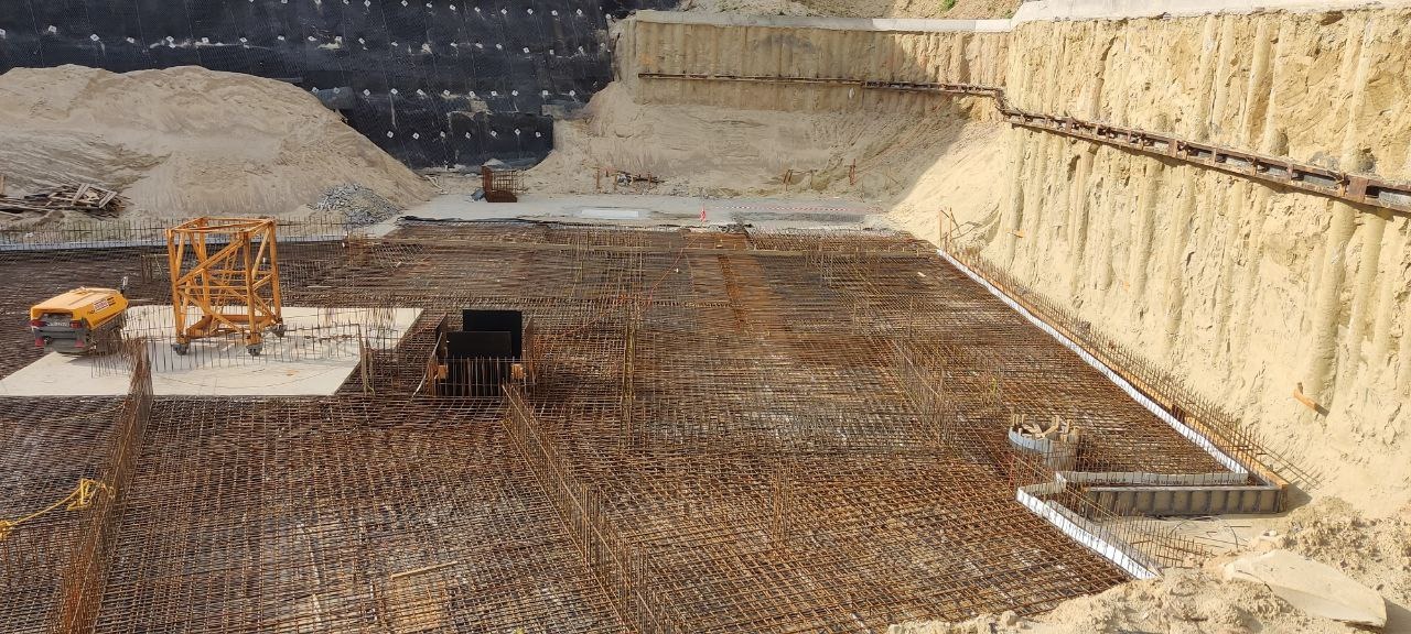 Szeroki kadr z placu budowy: zbrojenie fundamentów z widocznym wykopem, zabezpieczeniem ścian wykopu i sprzętem budowlanym. Widok z góry na siatkę zbrojeniową.