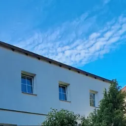 Biała elewacja budynku z trzema oknami w Ostródzie, widok z dołu na błękitne niebo z chmurami. Minimalistyczny styl, kontrastowe kolory.