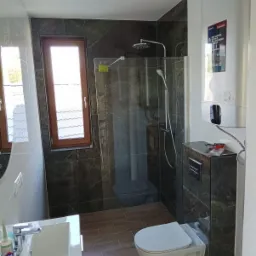 Nowoczesna łazienka z kabiną prysznicową typu walk-in, marmurowymi płytkami i podwieszaną toaletą. Widoczny bojler i okno z drewnianą ramą.