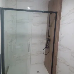 Kabina prysznicowa z czarną ramą i przesuwanymi drzwiami, zamontowana w łazience wyłożonej marmurkowymi płytkami, z drewnopodobnym panelem prysznicowym i czarną armaturą.