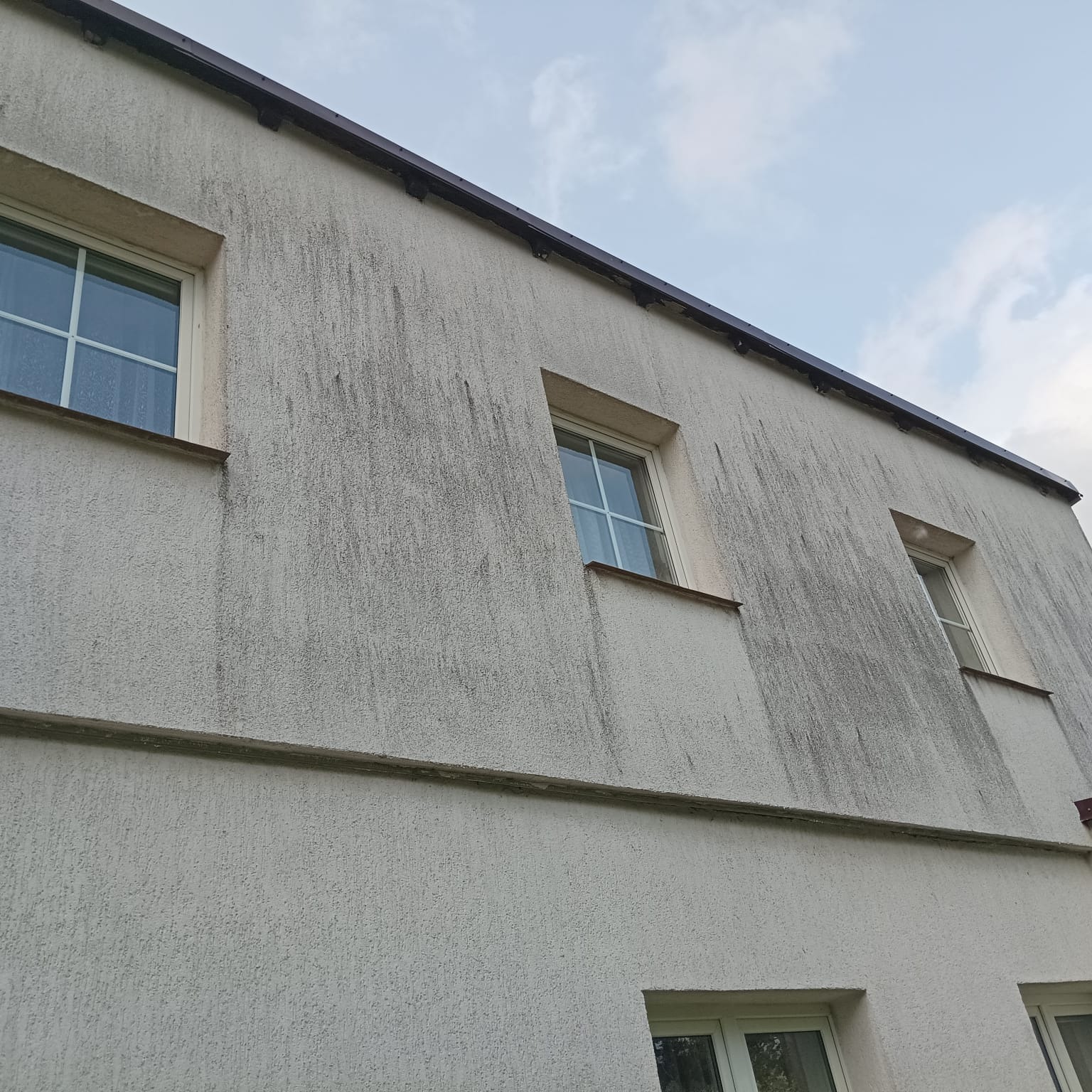 Brudna, biała elewacja budynku z trzema oknami na górze i dwoma na dole, widoczne zacieki i zabrudzenia. Perspektywa od dołu w kierunku nieba.