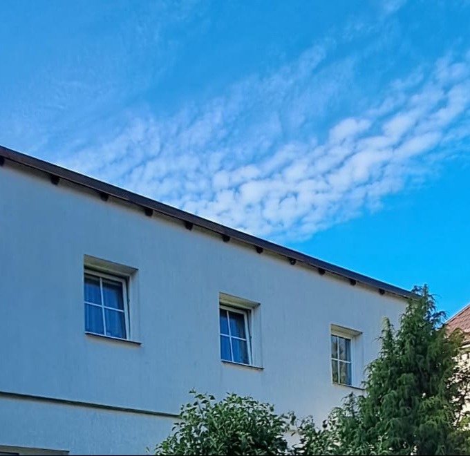 Biała elewacja budynku z trzema oknami w Ostródzie, widok z dołu na błękitne niebo z chmurami. Minimalistyczny styl, kontrastowe kolory.