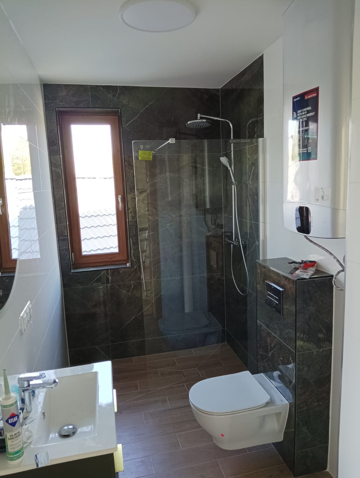 Nowoczesna łazienka z kabiną prysznicową typu walk-in, marmurowymi płytkami i podwieszaną toaletą. Widoczny bojler i okno z drewnianą ramą.